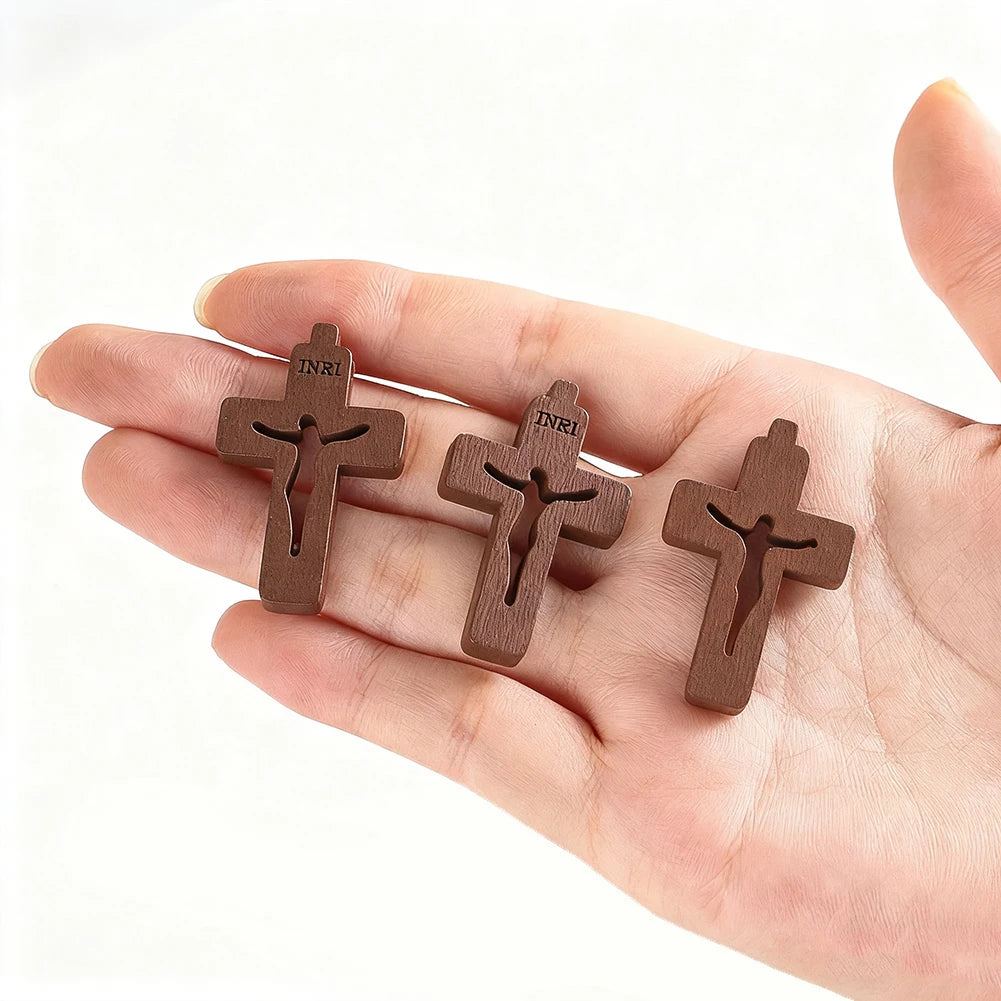 Trois petits pendentifs en bois de forme de croix sont tenus dans la main d'une personne sur un fond blanc.