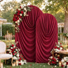 A deep red velvet drape serves as an arch cover for a wedding or event backdrop surrounded by floral arrangements.