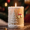 A white candle with a frosted design featuring a Christmas tree and golden bells, set against a blurred holiday background.
