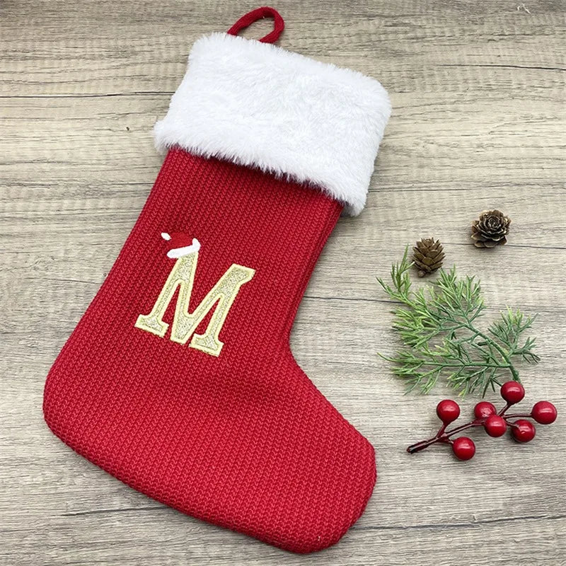 A red knitted sock with a white fluffy cuff and a gold letter M is displayed on a wooden surface.