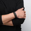 A close-up of a hand wearing a black braided leather cuff bracelet on a wrist, against a dark background.