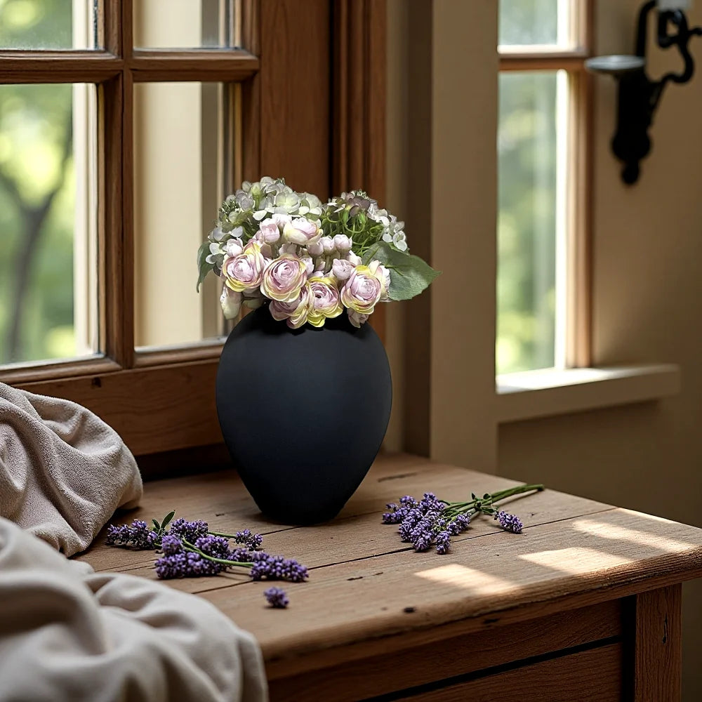 A dark gray modern round metal decorative vase filled with light pink and green floral arrangement sits on a wooden table nea
