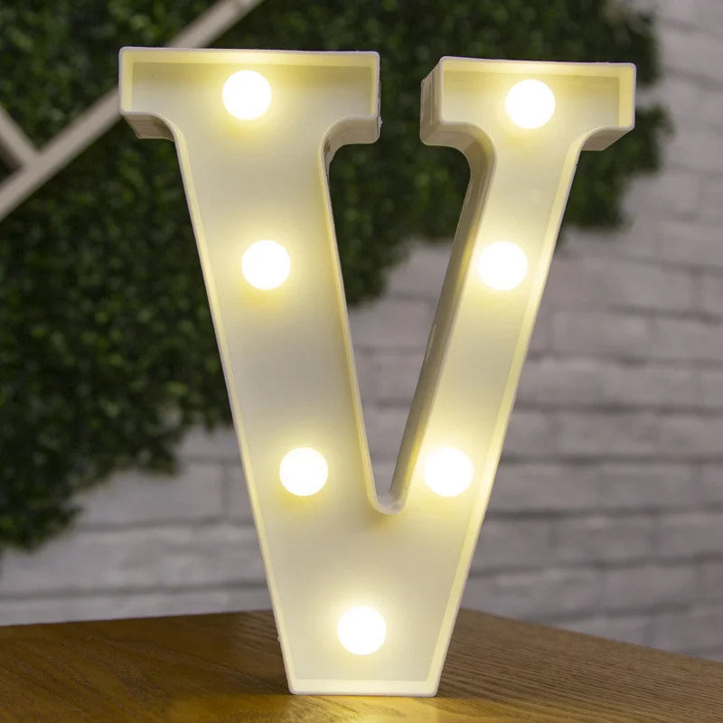 A large, illuminated letter V made of warm white LED lights standing on a wooden surface.