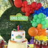 A display of red, orange, green, and blue latex balloons arranged in a cluster against a backdrop of green foliage.