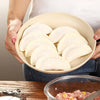 A hand holds a light-colored dumpling maker filled with white dumplings, next to a glass bowl of ground meat.