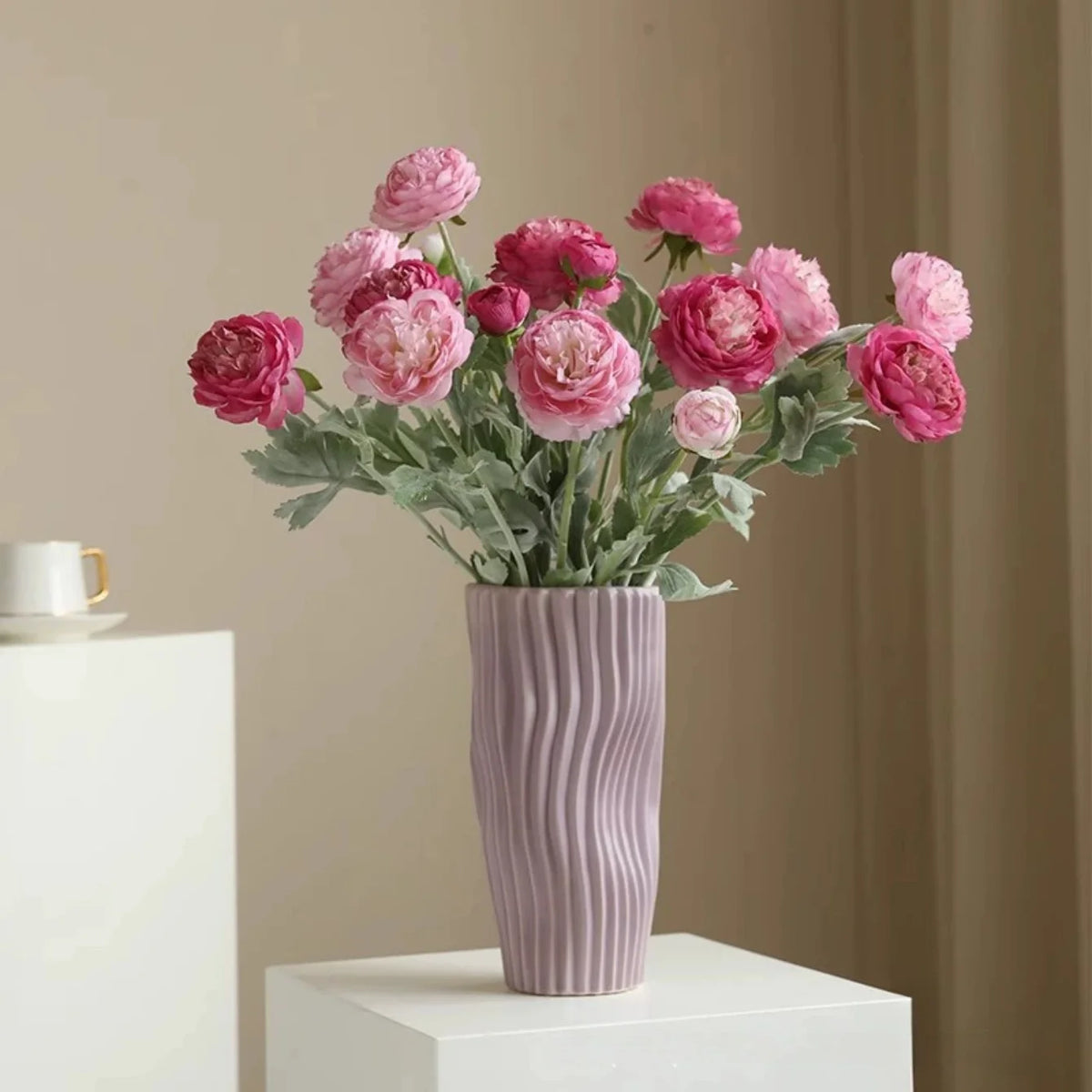 A bouquet of pink and magenta silk peony flowers in a ribbed lilac vase on a white surface.
