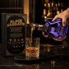 A hand pours whiskey from a bottle into a glass on a dark surface next to a Black Sabbath guitar-shaped decanter.