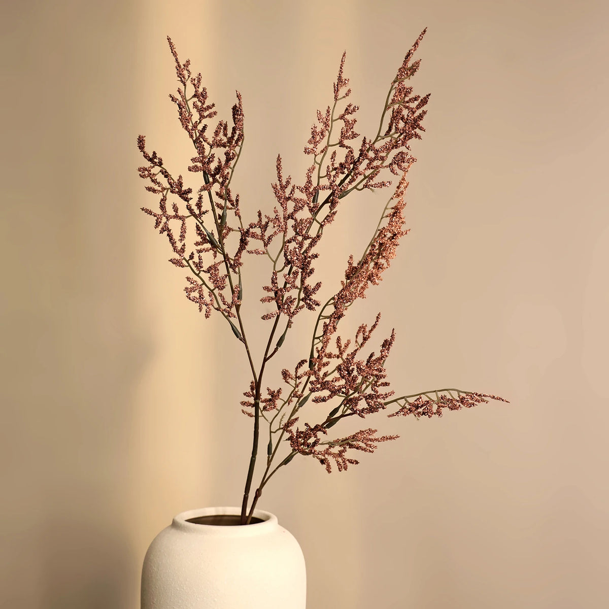 A dried branch of artificial silk lover grass in pinkish-red tones displayed in a white ceramic vase.