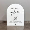 White acrylic sign with black script text that reads Let Love Glow and features an illustration of a glow stick.