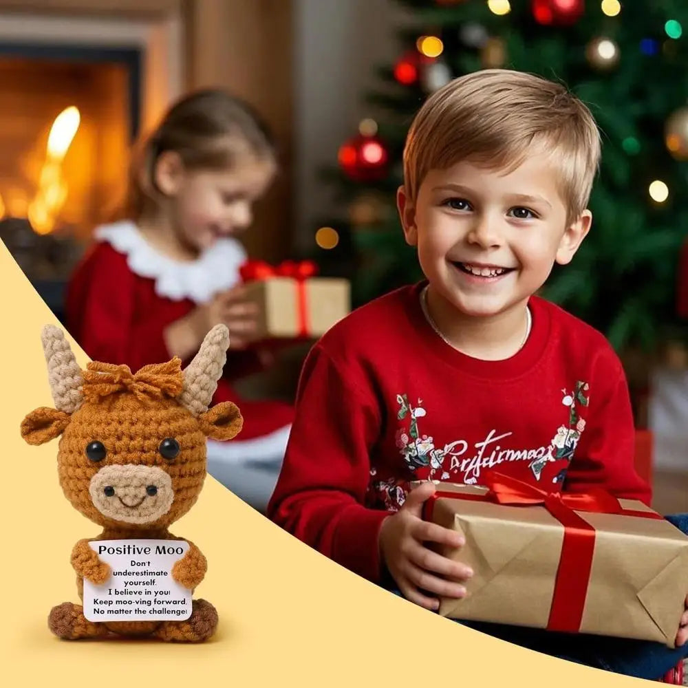A smiling boy holds a gift box in front of a Christmas tree with a crocheted Highland cow keychain keepsake.
