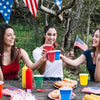Four women laughing outdoors holding red and blue plastic shot cups for a party event.