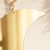 A gold moon-shaped acrylic wall mirror hanging by a chain, casting shadows on a light wall.