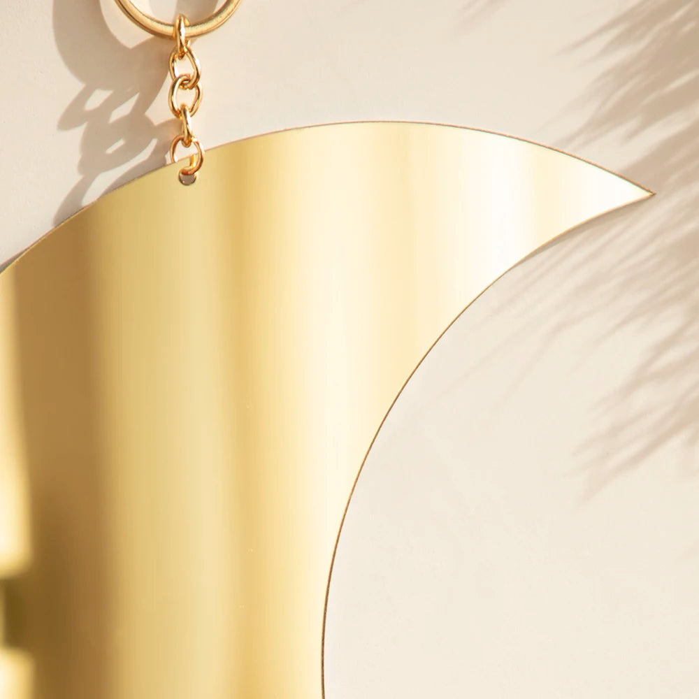 A gold moon-shaped acrylic wall mirror hanging by a chain, casting shadows on a light wall.