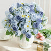 Un ramo de rosas y margaritas de seda artificiales azules y lavanda en un jarrón blanco sobre una mesa.