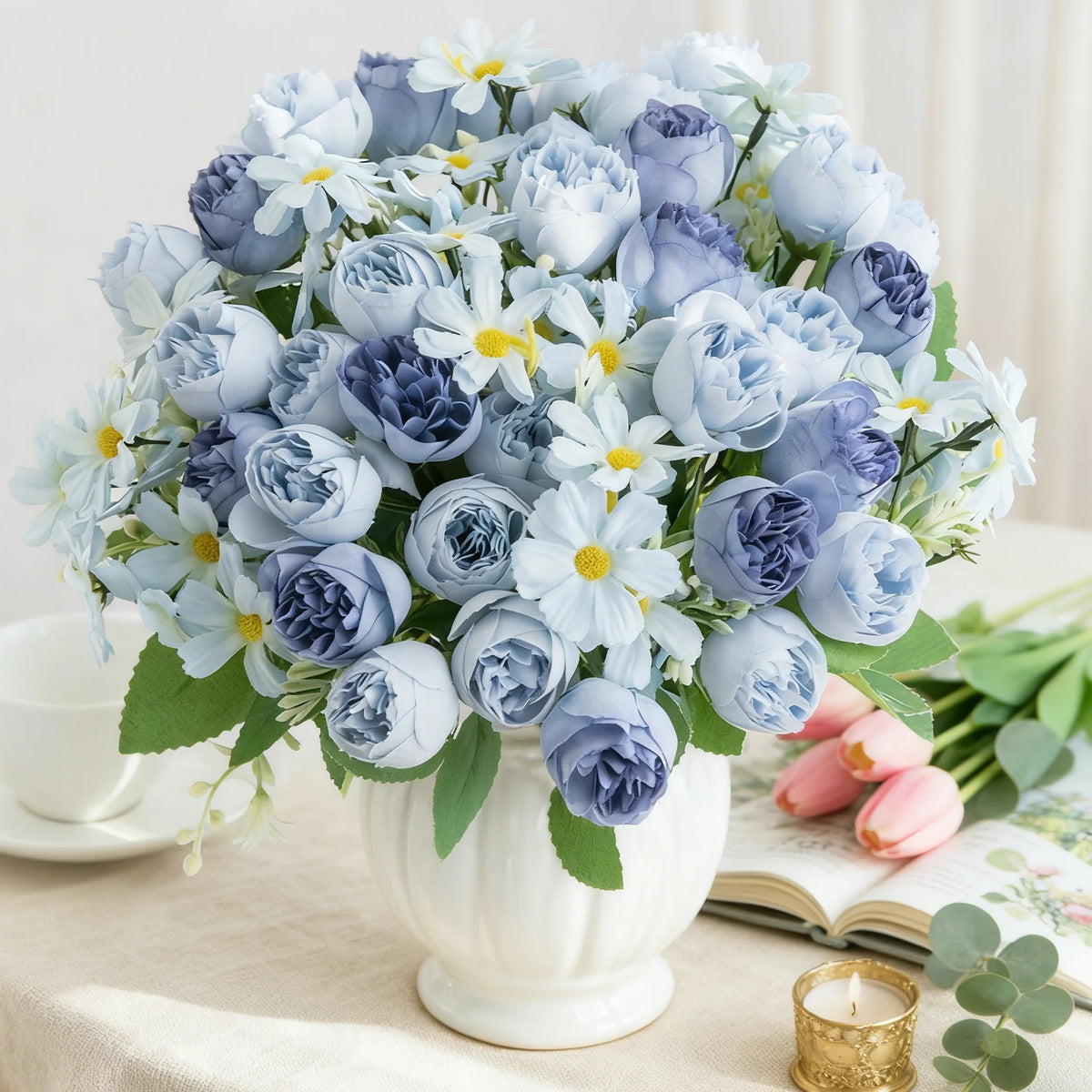 Un ramo de rosas y margaritas de seda artificiales azules y lavanda en un jarrón blanco sobre una mesa.