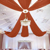 A wedding or event setup featuring a grand chandelier suspended beneath draped orange curtains and white sheer fabric.