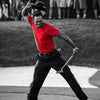 A man in a red shirt is swinging a golf club on a golf course, with spectators blurred in the background.