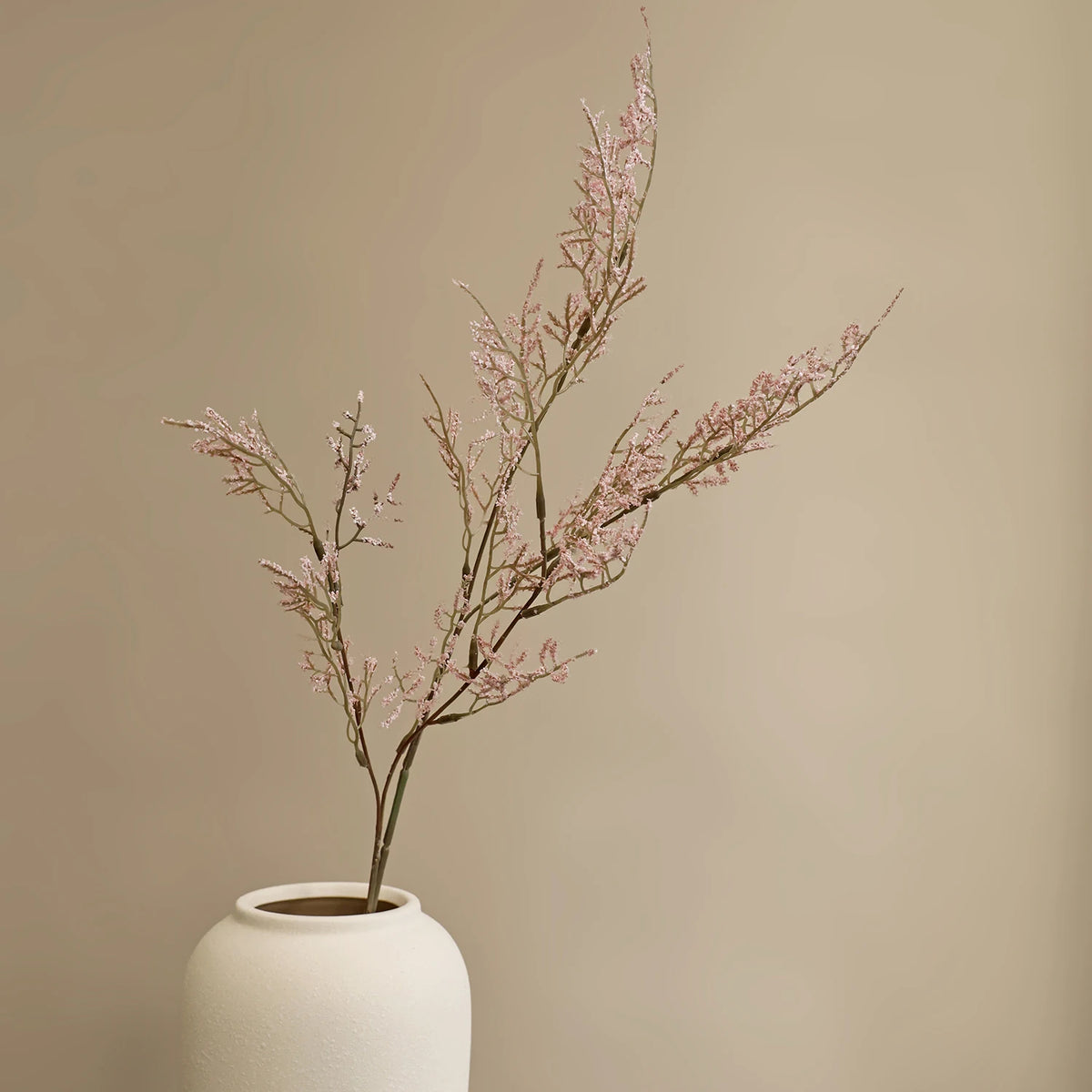 A slender branch of pink artificial silk lover grass displayed in a white ceramic vase against a neutral background.