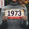 A person holds a vintage metal tin sign with the year 1973 printed on it, against a gray sweater.