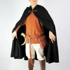 A male model wearing an orange tunic, white pants, and a black velvet hooded cloak with brown leather accents.