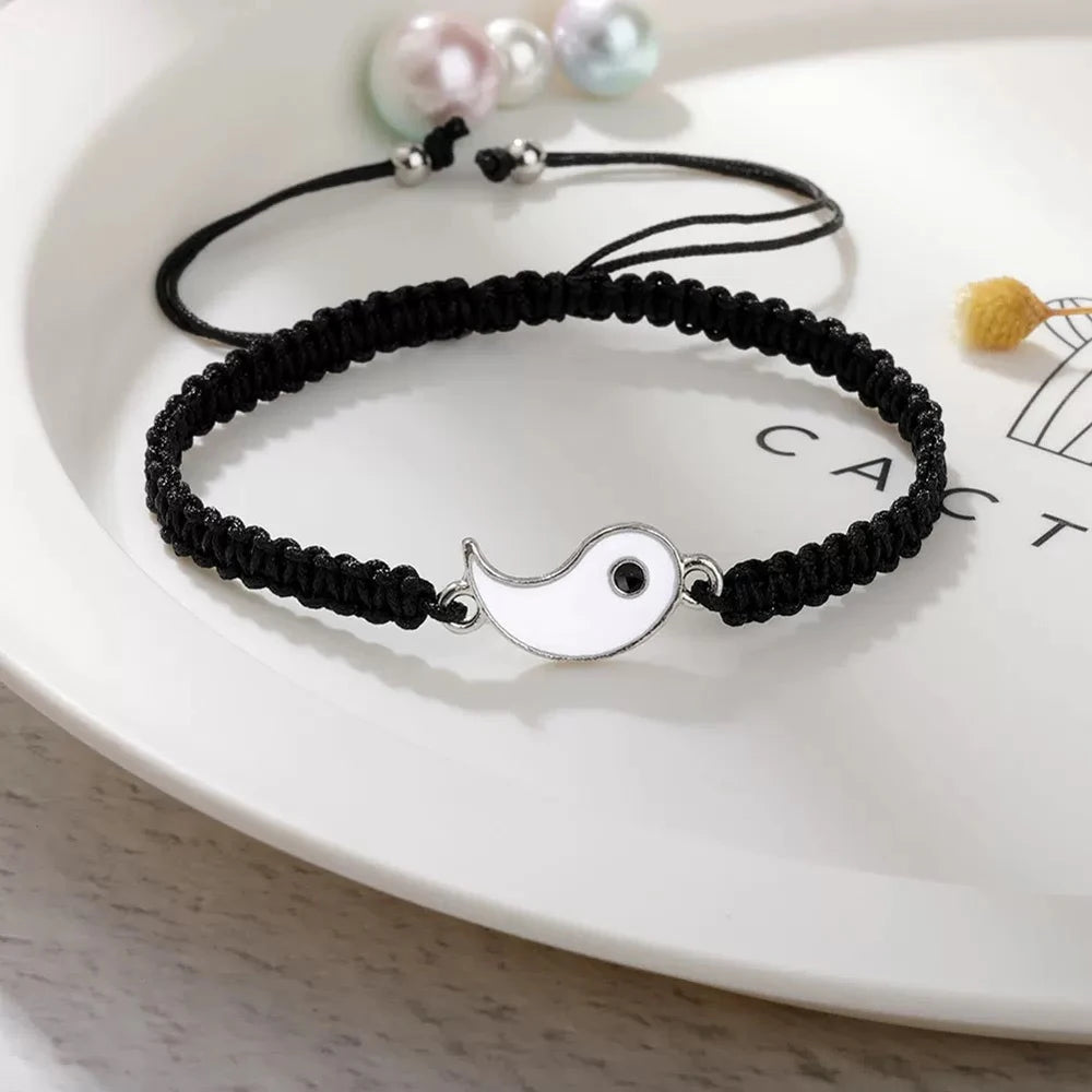 A black braided bracelet set featuring a silver yin-yang charm and pearl beads displayed on a white plate.