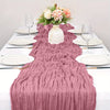 A pink boho style cheesecloth table runner draped over a white tablecloth with plates and a floral centerpiece.
