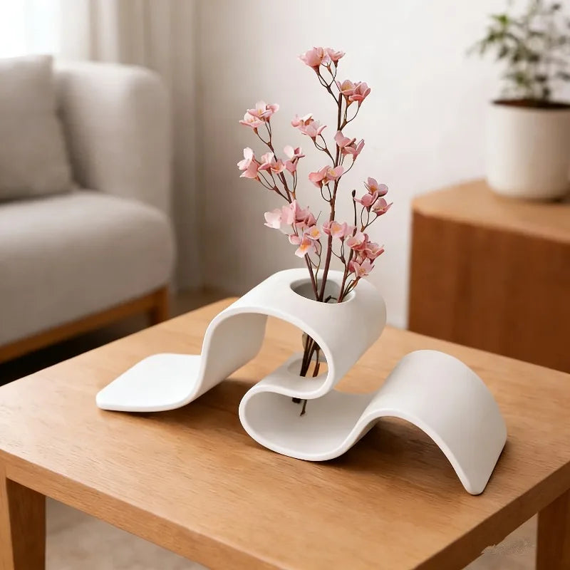 Jarrón blanco moderno curvo con ramas rosas sobre una mesa de madera.