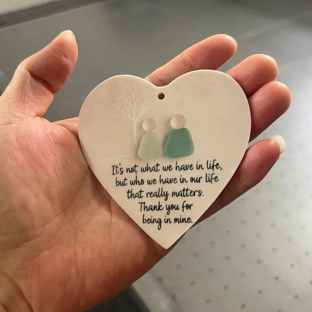 A hand holds a heart-shaped acrylic pendant featuring two small, light green figures.