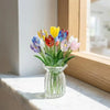 A bouquet of colorful glass flowers in a clear vase sits on a white marble surface next to a wooden frame.
