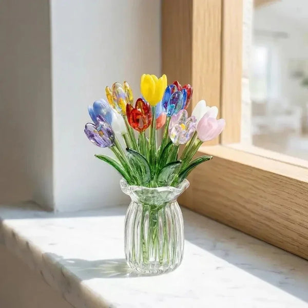 A bouquet of colorful glass flowers in a clear vase sits on a white marble surface next to a wooden frame.
