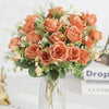 A bouquet of five peach-colored silk artificial roses with green foliage displayed in a clear glass vase.