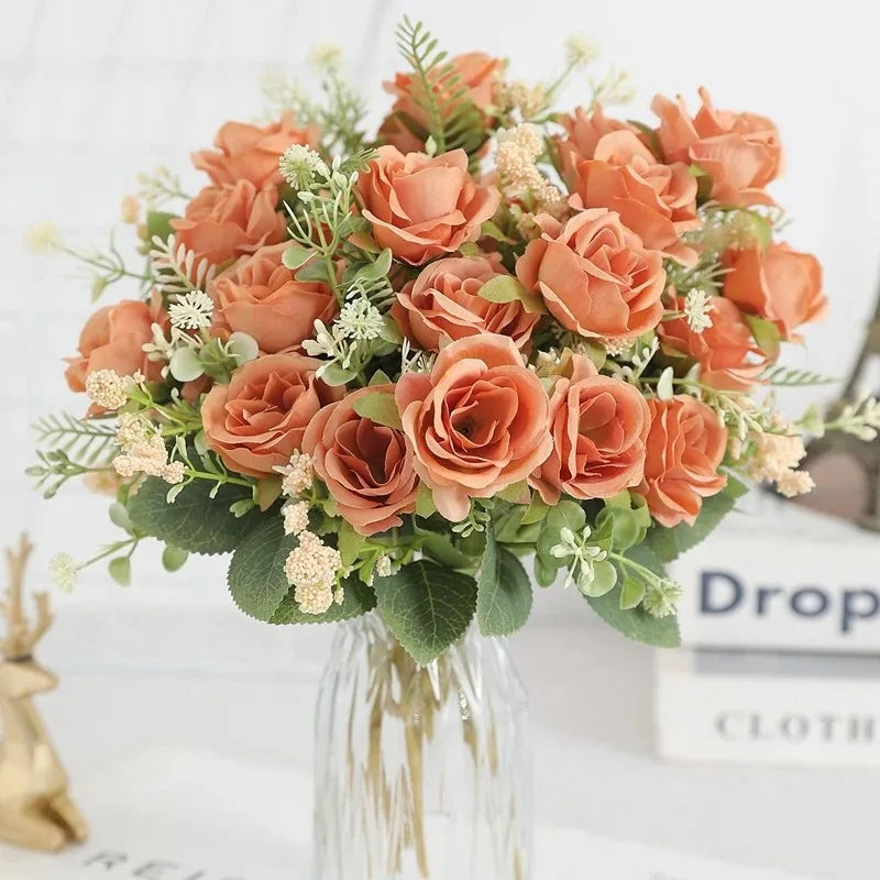 A bouquet of five peach-colored silk artificial roses with green foliage displayed in a clear glass vase.