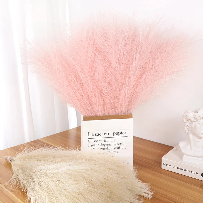 A bouquet of pink faux pampas grass reeds displayed in a cardboard box on a wooden surface.