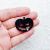 Una mano sostiene un colgante de acrílico negro de Halloween con la cara de una calabaza tallada y la silueta de un gato pequ