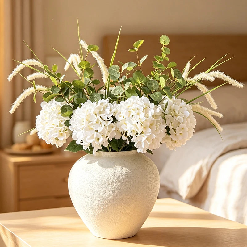 A rustic textured round white vase filled with green and cream foliage sits on a wooden surface.