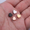 A hand holds several heart-shaped stainless steel bead charms in silver, black, and gold finishes.