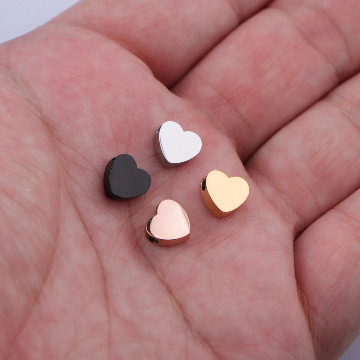 A hand holds several heart-shaped stainless steel bead charms in silver, black, and gold finishes.