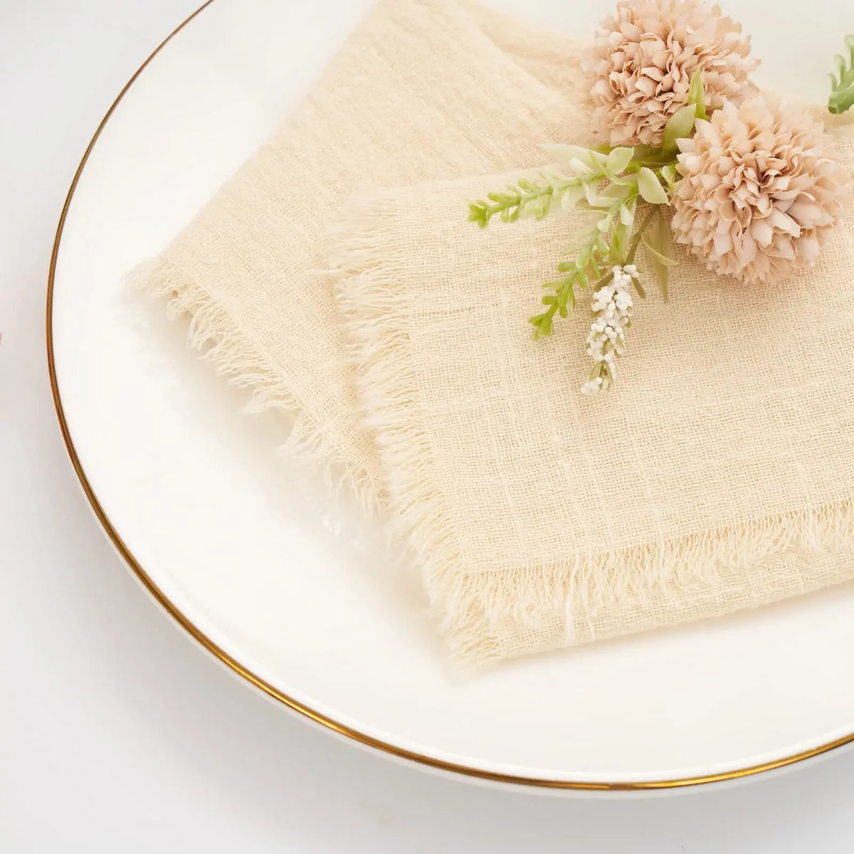 A set of ten handcrafted fringed cotton napkins in natural beige with dried floral accents on a white plate.
