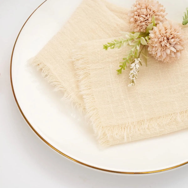 A set of ten handcrafted fringed cotton napkins in natural beige with dried floral accents on a white plate.