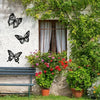 Three black metal butterfly wall art pieces displayed on a white stucco wall next to a window and green foliage.