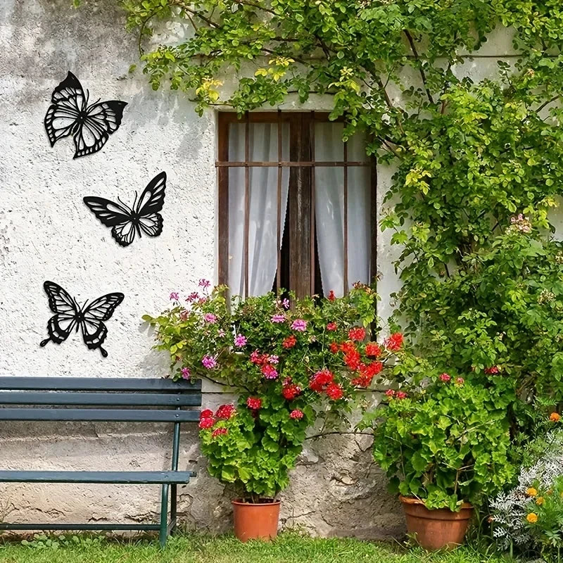Three black metal butterfly wall art pieces displayed on a white stucco wall next to a window and green foliage.