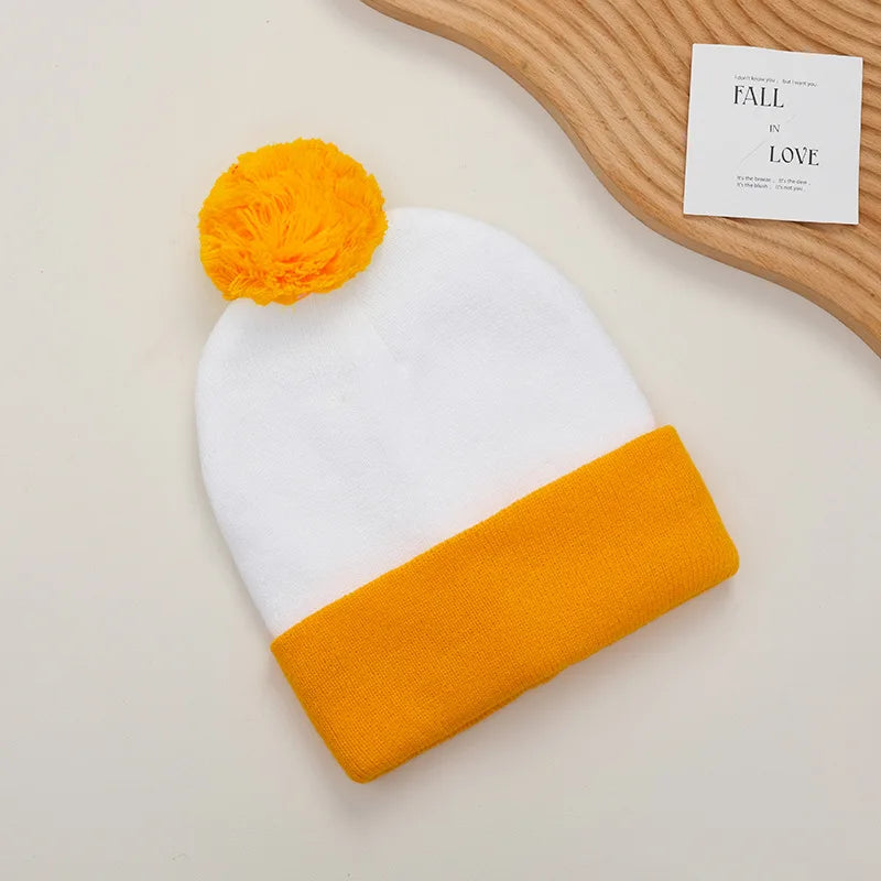 A knitted beanie in white with an orange pom-pom and an orange cuff, displayed next to a wooden surface.