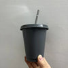 A hand holds a dark gray plastic cold cup with a lid and straw against a light gray wall.