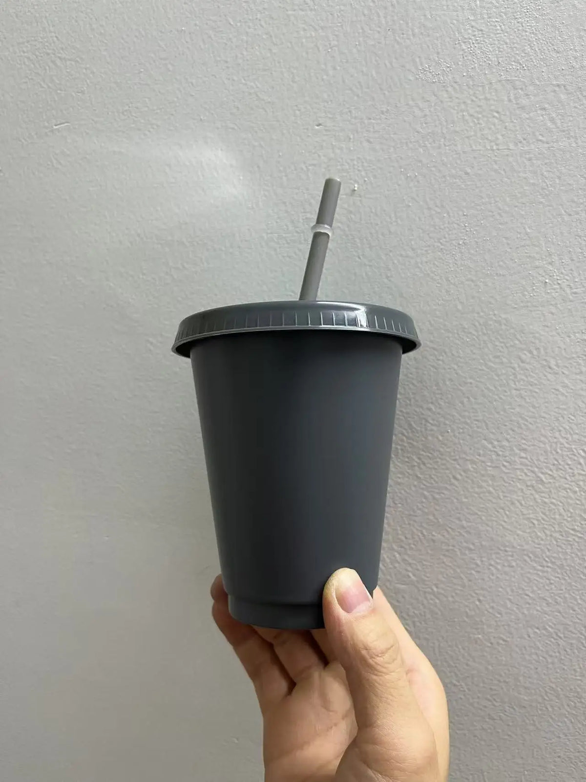 A hand holds a dark gray plastic cold cup with a lid and straw against a light gray wall.