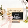 A wooden ultrasound photo frame with a black screen displaying an ultrasound image and a pink ribbon for a pregnancy announce