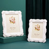 A white ornate photo frame with a cream mat displaying a pink rose floral design, set against a dark teal background.