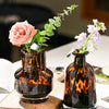 A dark amber glass vase holds a pink rose, displayed on a wooden board.