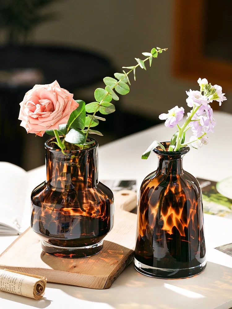 A dark amber glass vase holds a pink rose, displayed on a wooden board.