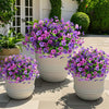 A collection of vibrant purple and pink artificial flowers planted in white pots on a paved outdoor surface.