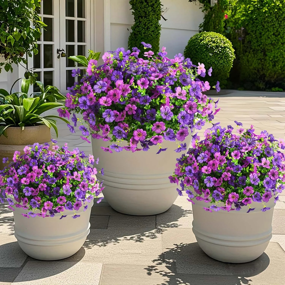 A collection of vibrant purple and pink artificial flowers planted in white pots on a paved outdoor surface.